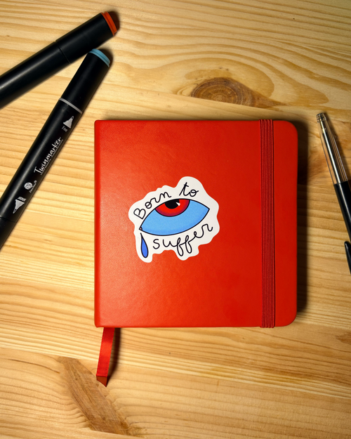 Sticker « Born to suffer » avec un œil bleu et une larme, collé sur un carnet rouge posé sur une table en bois.