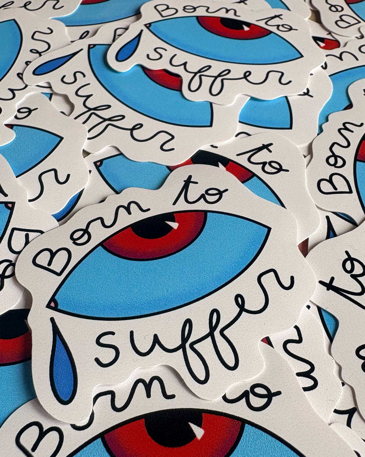 Stickers « Born to suffer » superposés, représentant un œil bleu à l’iris rouge, une larme et du texte noir.