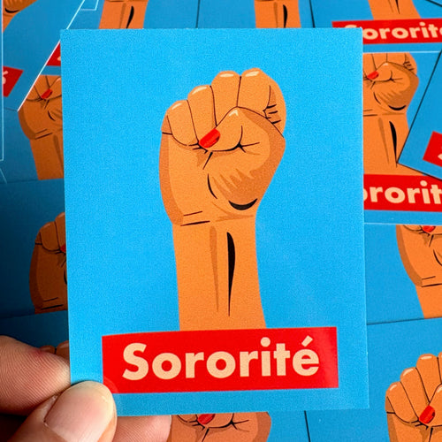 Sticker « Sororité » tenu à la main, avec un poing levé et un ongle rouge sur fond bleu.
